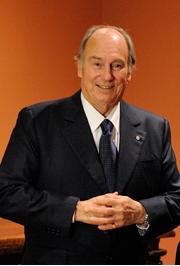 His Highness Prince Karim Aga Khan smiling while standing in a suit, showcasing a welcoming gesture.