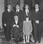Historical black-and-white photograph of His Highness Prince Karim Aga Khan seated in a wheelchair, surrounded by five young men in suits and a young girl in a coat.