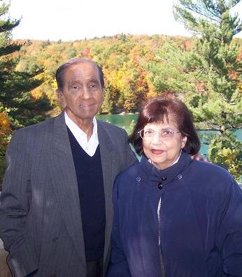 An older couple standing outdoors near a lake surrounded by trees in vibrant autumn colors.