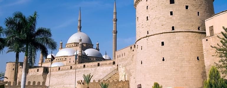 A panoramic view of a historic mosque and fortress, showcasing intricate architectural features and palm trees in the foreground under a clear blue sky.