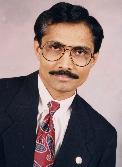 A man wearing glasses and a formal black suit with a patterned tie, posed against a neutral background.