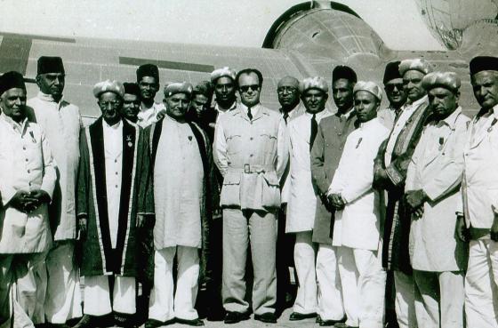 Prince Aly Khan with members of the Ismaili community in front of an airplane.