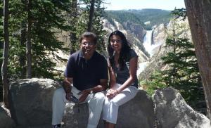 Nurin Merchant and her father sitting on a rock in Yellowstone National Park, with a waterfall and trees in the background.