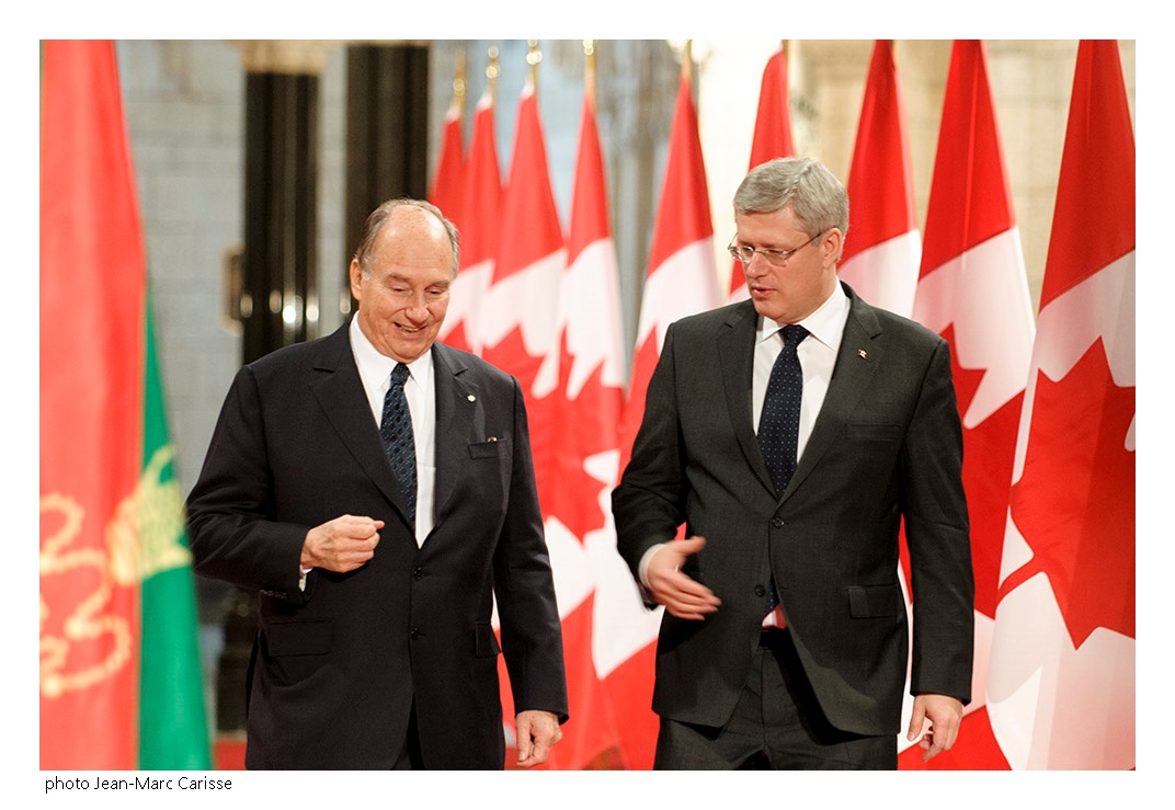 His Highness the Aga Khan and Prime Minister Harper in a conversation as they proceed to the signing ceremony of  the protocol of understanding between the Ismaili Imamat and Canada. They are flanked on either side by the flags of the red and green flags of the Ismaili Imamat and the maple leaf of Canada. Photo: Jean-Marc Carisse. Copyright.