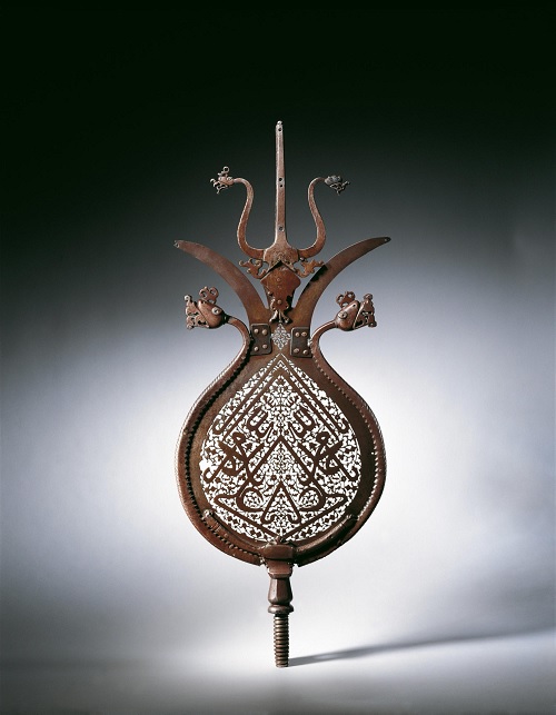Processional standards (‘alams) are used in Shia processions, particularly on the day of ‘Ashura, the tenth day of the month of Muharram, to commemorate the martyrdom of the Prophet Muhammad’s grandson, al-Hosayn, the son of ‘Ali, in the seventh century in Karbala’, Iraq. In this openwork ‘alam, the form, decorative elements, and function are closely intertwined. The bifurcated blades on the top of the pear-shaped body of this beautifully carved ‘alam are a symbolic reference to the first Shia imam, ‘Ali, the Prophet’s son-in-law, who is known by the epithet dhu’l-fiqar in reference to his bifurcated sword. ‘Ali is also referred to by name in the mirror-image inscription on the central field of this ‘alam: ya Allah ya Muhammad ya ‘Ali, calling upon God, Muhammad, and ‘Ali for support. The symmetrical formation of the invocation ya ‘Ali in the inscription is usually seen as depicting the stylized face of a lion, another symbolic reference to the first imam. Photo and caption: Aga Khan Museum. Accession Number: AKM679, 82cm x 32.5 sm, Iran or India,16th Century, Pierced Steel.