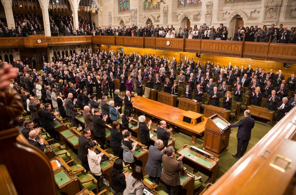 A large audience, including Ismaili leaders and Canadian politicians, stands and applauds in a formal legislative chamber during a significant event recognizing His Highness the Aga Khan on his anniversary as the Ismaili Imam.