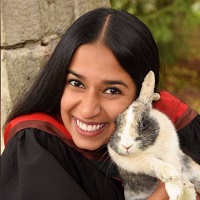 A woman wearing a graduation gown and hood smiles while holding a rabbit against a natural backdrop.