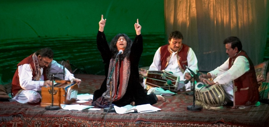 Abida Parveen performing on stage with three musicians, passionately singing with her hand raised, surrounded by traditional instruments.