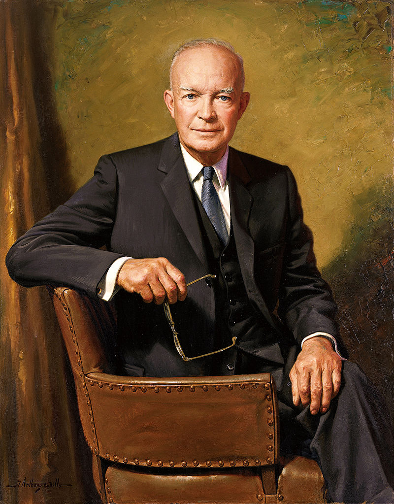 A portrait of President Dwight D. Eisenhower, seated in a brown leather chair, holding a pair of glasses, wearing a dark suit and tie, with a neutral background.