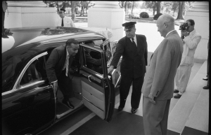 Historic black and white photo showing a man exiting a car while a uniformed officer assists him, with another man, presumably Dwight D. Eisenhower, standing nearby.