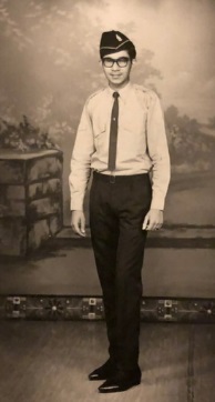 A young man in a formal uniform and glasses stands posed for a portrait against a painted backdrop, showing an outdoor scene.