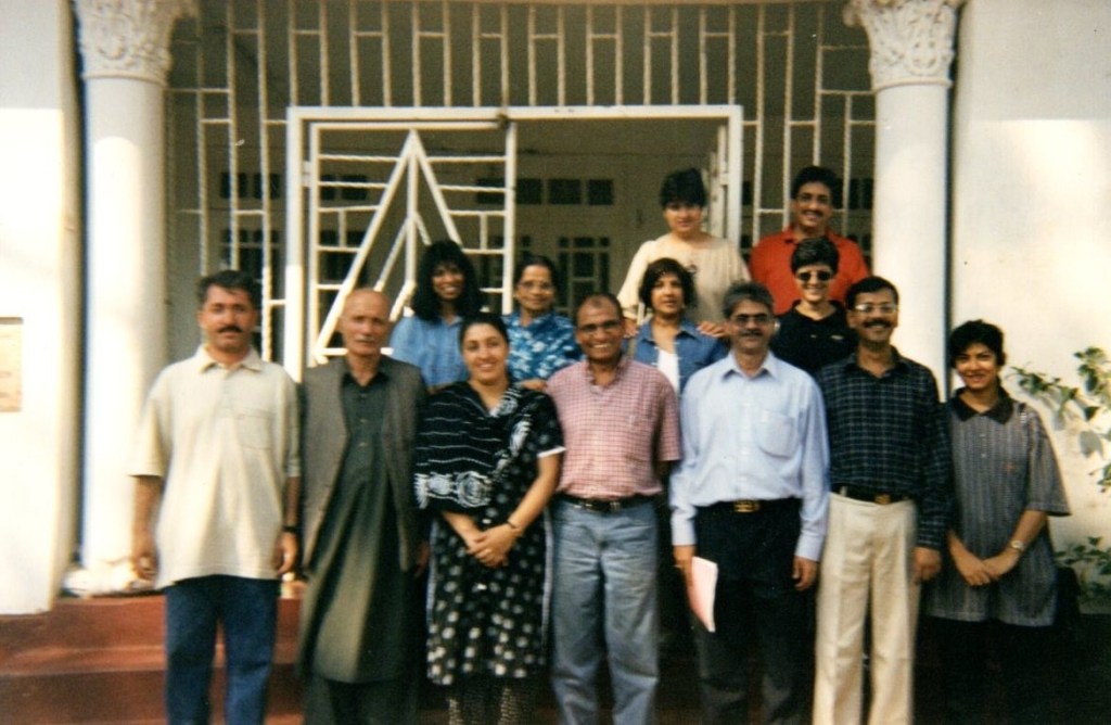 Group photo of individuals standing and sitting in front of a decorative building, showcasing a mix of adults and children, all smiling and posing together.