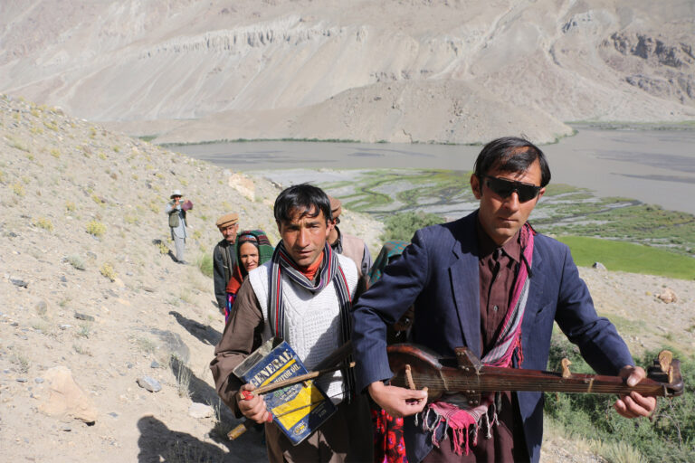 A group of people trekking along a mountainside near the Amu Darya river, with one person holding a traditional string instrument, while others carry items and wear traditional clothing.