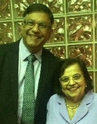 A man and a woman smiling together, standing close to each other in front of a glass brick wall.