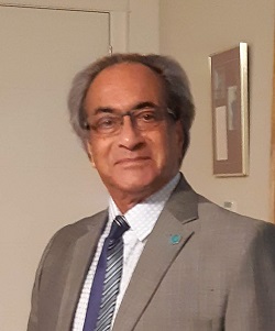 A man in a suit stands in a room with a neutral-colored wall and a door in the background, smiling at the camera.