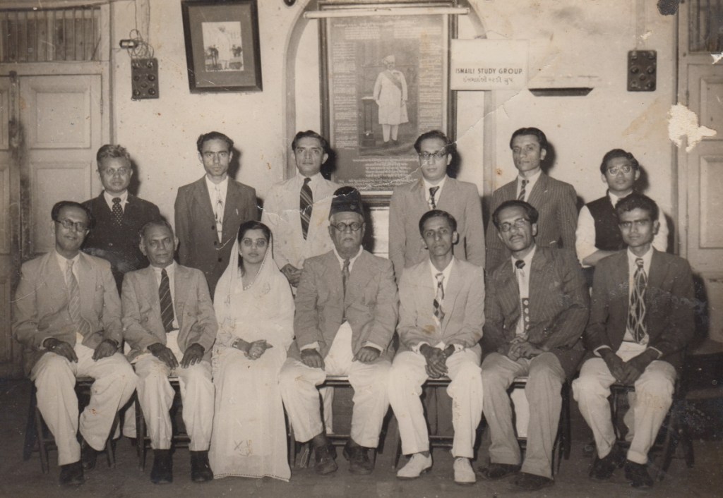 A black and white historical photo featuring a group of people, including men in suits and a woman in traditional attire, posing together in front of a wall with framed pictures and a sign that says 'Ismaili Study Group.'