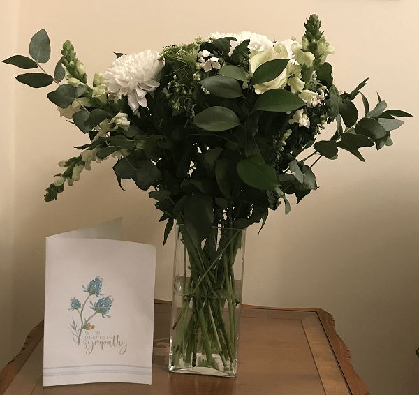 A bouquet of white and green flowers in a clear glass vase, placed next to a sympathy card on a wooden table.