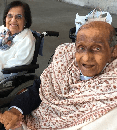 An elderly couple smiling and sitting together in wheelchairs, sharing a moment of affection.