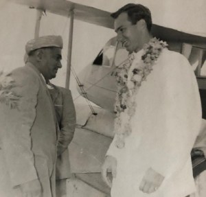 Historical photograph of two individuals, one wearing a traditional outfit and the other in a formal attire with a garland, engaged in conversation near an aircraft.