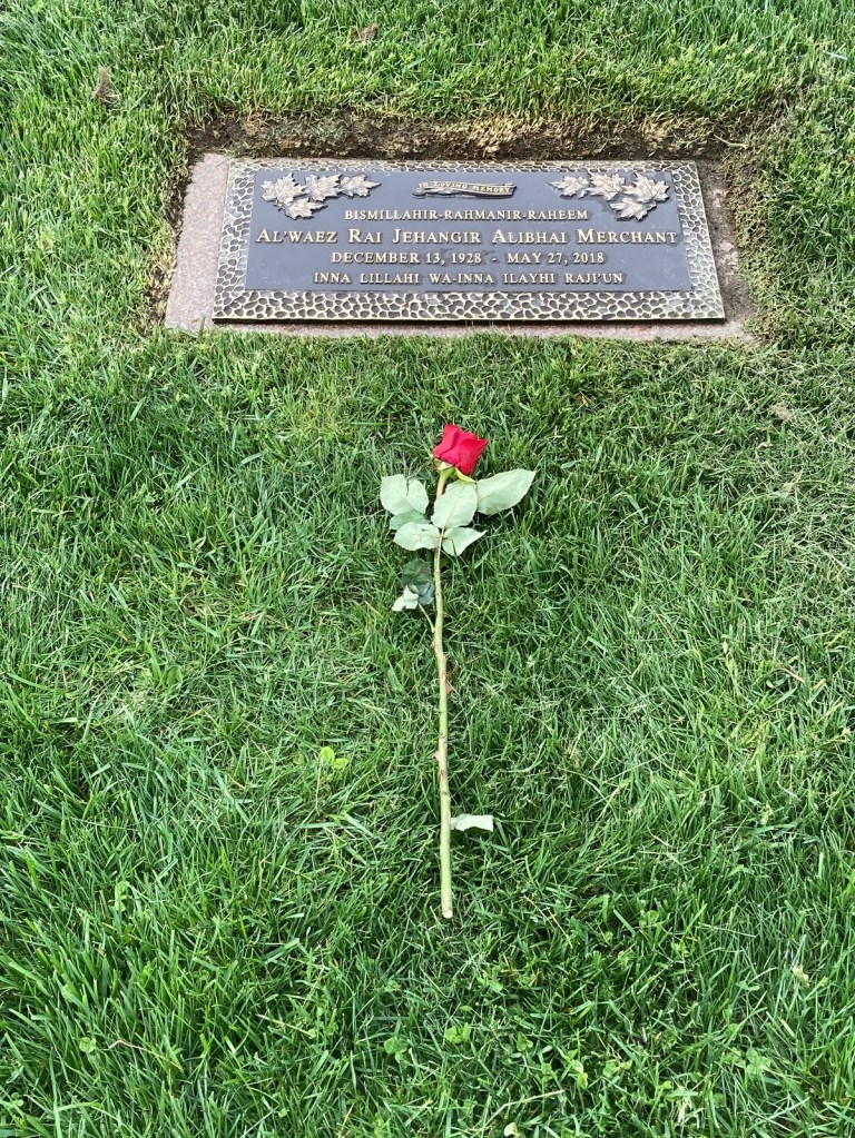 A gravestone with an engraved plaque memorializing Al'waez Rai Jehangir Alibhai Merchant, dated December 13, 1928 – May 27, 2018, placed in green grass with a red rose resting beside it.