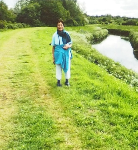 A woman standing on a grassy path beside a river, wearing a blue shawl and white pants.