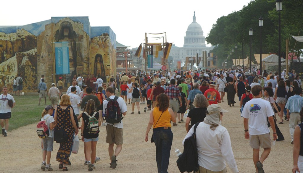 A large crowd walking on a pathway at an outdoor event, with decorative structures and the U.S. Capitol building in the background.