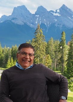 A smiling man sitting outdoors in front of a mountain landscape, surrounded by trees.