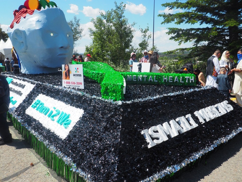 A colorful float representing mental health awareness features a large blue head sculpture and decorative elements, with text promoting Ismaili Muslims and community support.