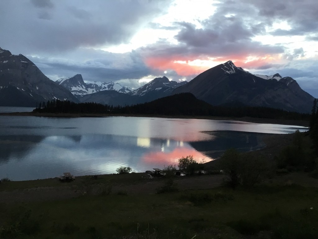 A stunning sunset over a tranquil lake, surrounded by the majestic Kananaskis Mountain Range in Alberta, Canada, reflecting colorful clouds in the water.