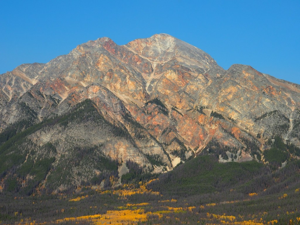 A vibrant view of Pyramid Mountain with colorful rock layers and a clear blue sky, surrounded by lush green forests and patches of autumn foliage.