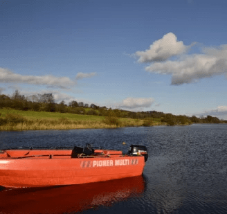 A red fishing boat named 'PIONER MULTI' anchored on a calm lake, surrounded by green hills and a blue sky with scattered clouds.