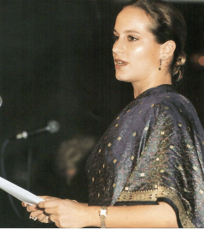 A woman speaking at an event, wearing a dark purple outfit with intricate designs, holding a piece of paper.