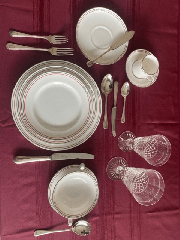 A beautifully arranged dining table setting featuring white plates with red rim, silver cutlery, and elegant glassware, all placed on a rich burgundy tablecloth.