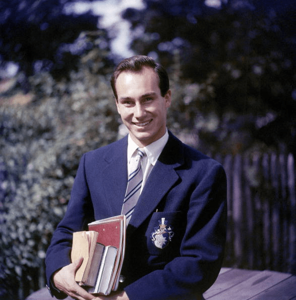 A young man smiling while holding a stack of books, dressed in a formal blazer with an emblem, standing outside in a lush green setting.