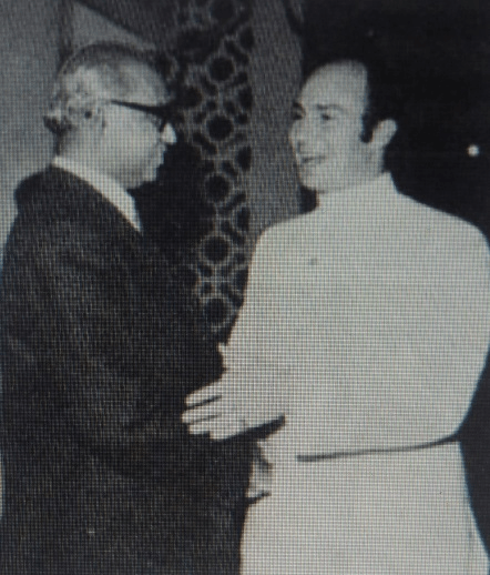 Two men engaged in conversation, one in a suit and glasses, the other in a white traditional outfit.