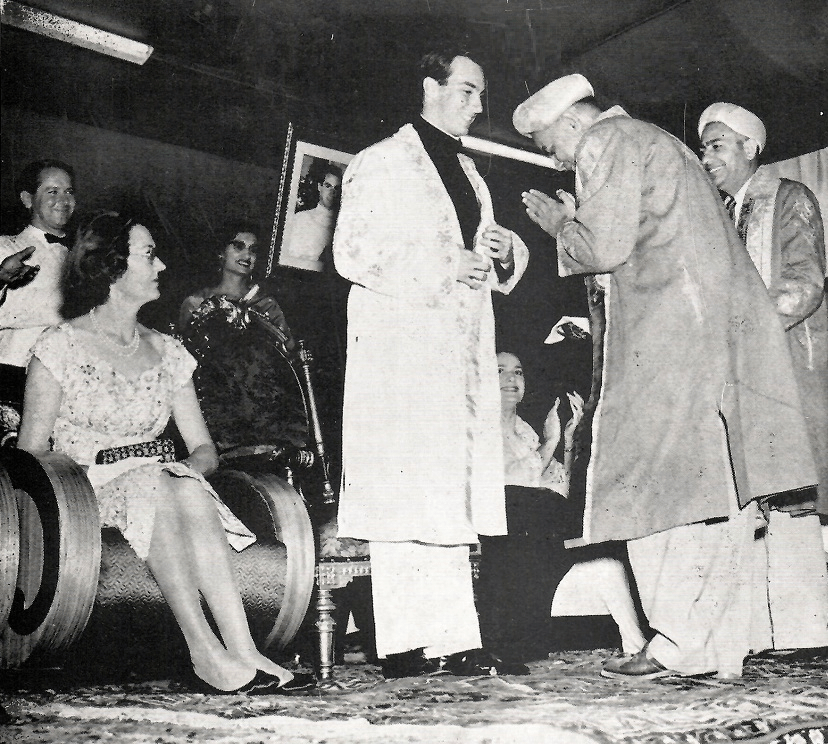 A historical black-and-white photo featuring a gathering with a woman in a dress sitting on a chair, while a man in traditional attire interacts with another man who is bowing, in a festive setting.