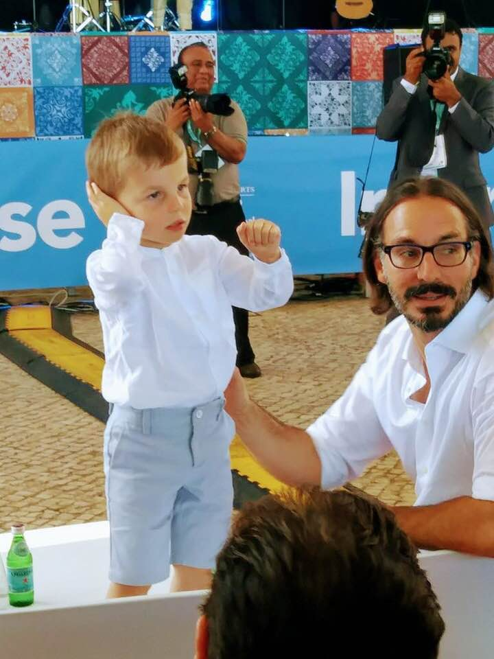 A young boy wearing a white shirt and light blue shorts stands with his hands on his ears, while a man in a white shirt looks at him. In the background, several photographers are capturing the moment, and there is a colorful backdrop.