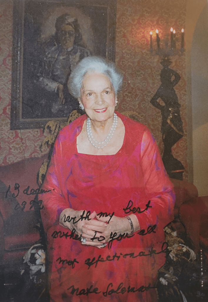 A woman in a red dress stands smiling in front of a decorated wall, with candles and a painting in the background, holding a handwritten note.