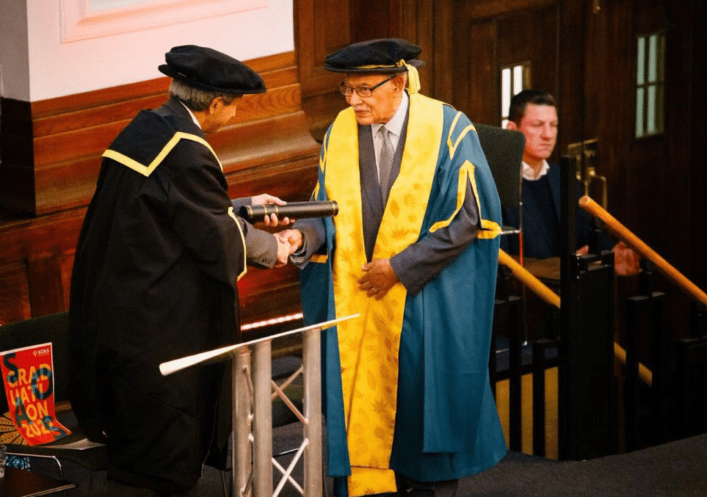 A formal graduation ceremony where a man in academic robes receives a diploma from another official, with guests seated in the background.