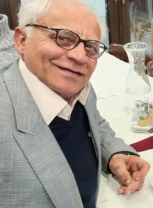 A portrait of an elderly man with glasses, smiling warmly at the camera, wearing a light grey blazer over a dark sweater.