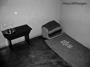 Black and white image of a small, sparsely furnished room containing a bedroll and a table with a bowl.