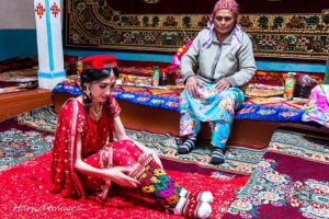 A woman in traditional attire sitting on a colorful rug, putting on a decorative leg accessory, while an older man in traditional clothing watches her.