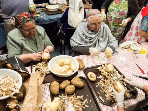 Two elderly women peeling potatoes at a communal kitchen table, surrounded by unpeeled and peeled potatoes.