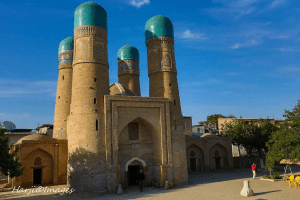 A historical building with four turquoise domes against a blue sky, situated in a courtyard with a person in the foreground.