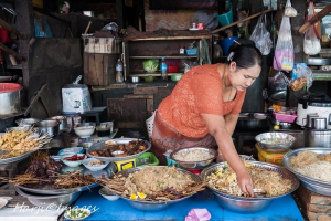 A woman in an orange shirt preparing street food in a bustling market, surrounded by bowls of various dishes and ingredients.