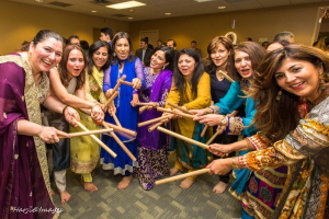 A group of women dressed in colorful traditional outfits joyfully participate in a festive dance, holding sticks in a circle.