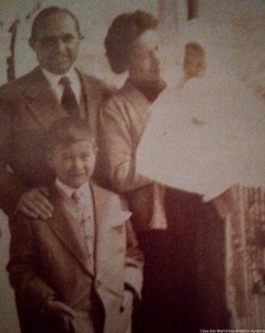 A sepia-toned family photograph featuring a man and a woman standing together, with a young boy in front of them. The man is smiling and wearing a suit, while the woman has her hair styled and is holding a white object, possibly a blanket or a dress. The boy is dressed in formal attire and looking at the camera.