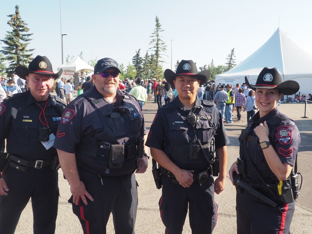 A group of four Calgary police officers posing together at the Ismaili Muslim Stampede Breakfast event, held outdoors with a crowd in the background.