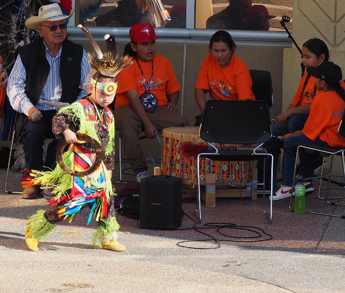 A young child wearing a colorful traditional outfit performs during a cultural event, while an audience of children in orange shirts and adults sits nearby, engaging with drums and musical instruments.