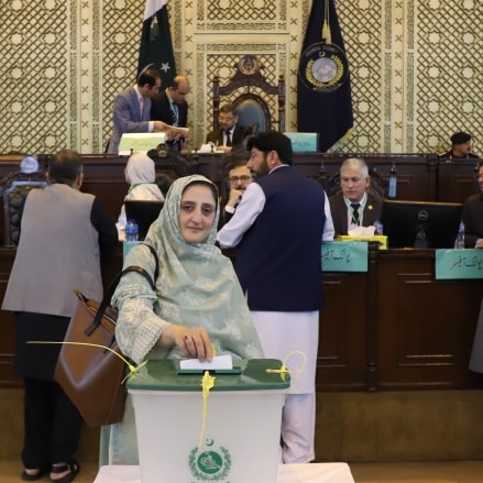 A woman in traditional clothing casting her vote at an election booth in a government assembly, surrounded by other officials and a large ornate backdrop.